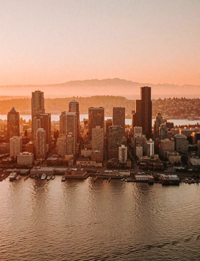 Seattle skyline at sunset seen across Elliott Bay with warm-lit towers and a distant mountain silhouette