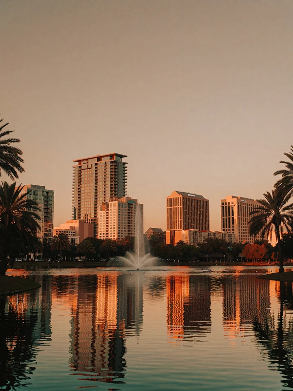 Downtown Orlando skyline reflected in a calm lake at dusk with a fountain and palm trees
